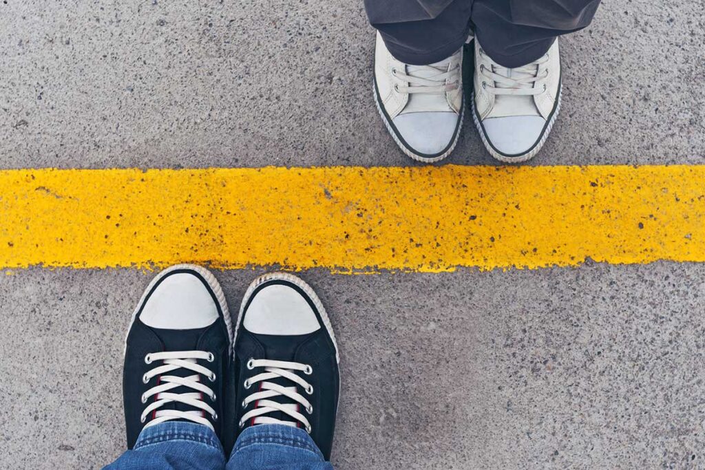 Embrace setting boundaries, overhead shot showing a line painted on asphalt with people in a set of sneakers standing on opposite sides of the line to represent boundaries